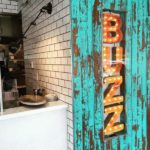 A marquee sign that reads BUZZ installed into a bark textured wood wall beside a white tiled commercial kitchen and serving area.