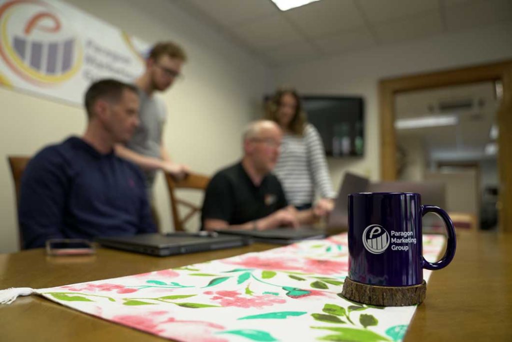 Close up of the Paragon Marketing team at a desk, with the focus on a coffee mug. Consider Paragon's marketing solutions for your business.